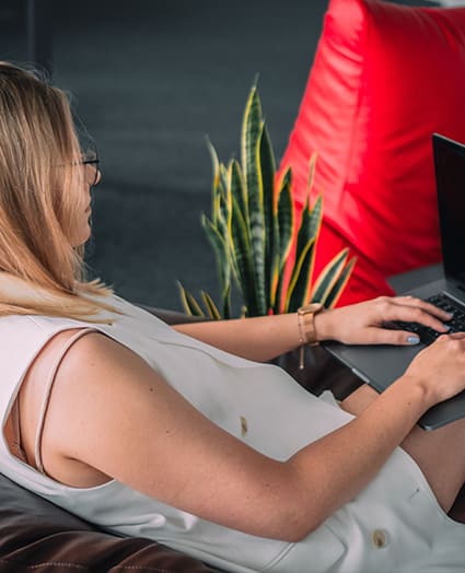 Kobieta siedzi na pufie bokiem do aparatu, trzymając na kolanach laptopa.